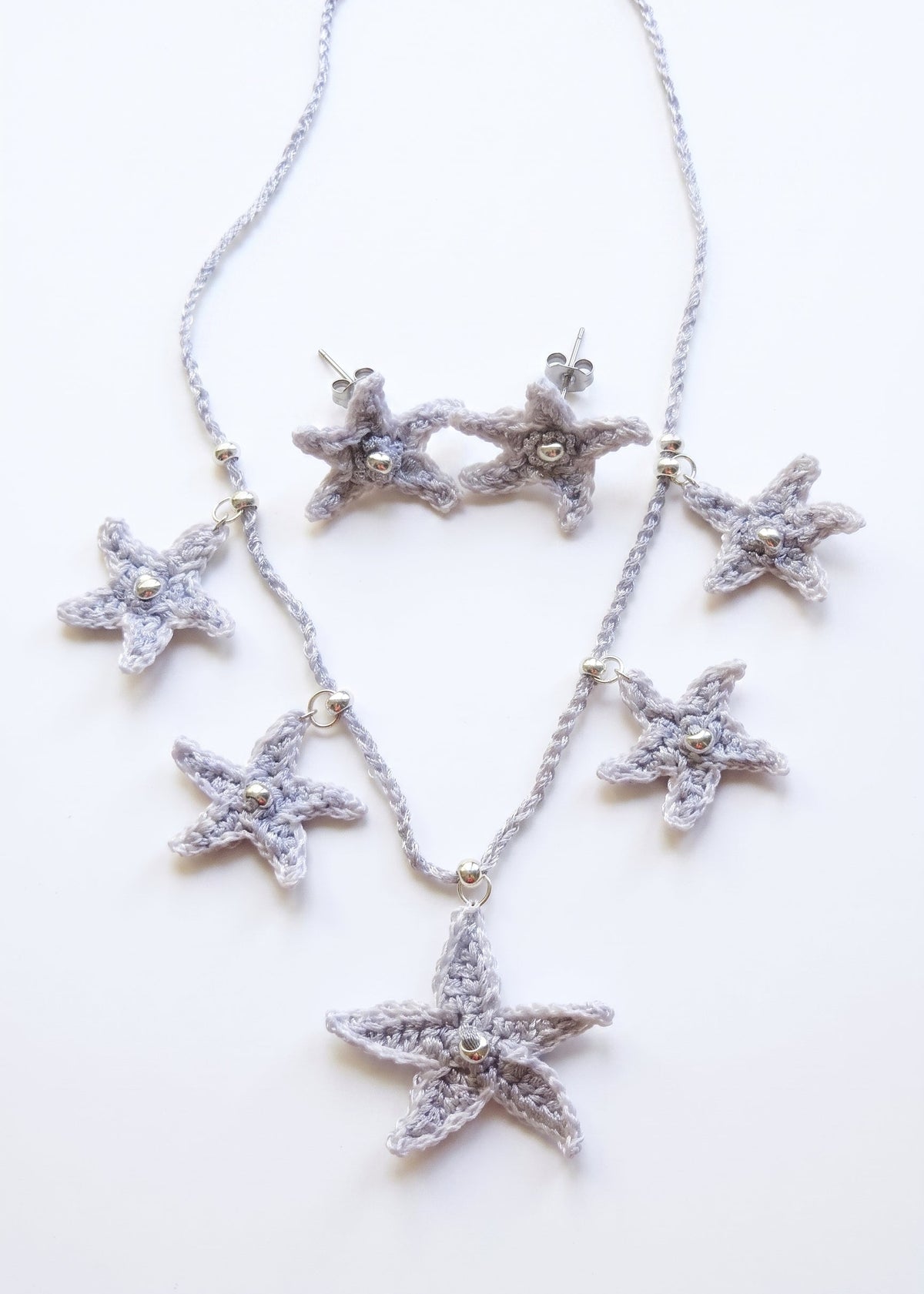Monográfico de conjunto de collar y pendientes de crochet con forma de estrellas de mar plateadas sobre base de madera.